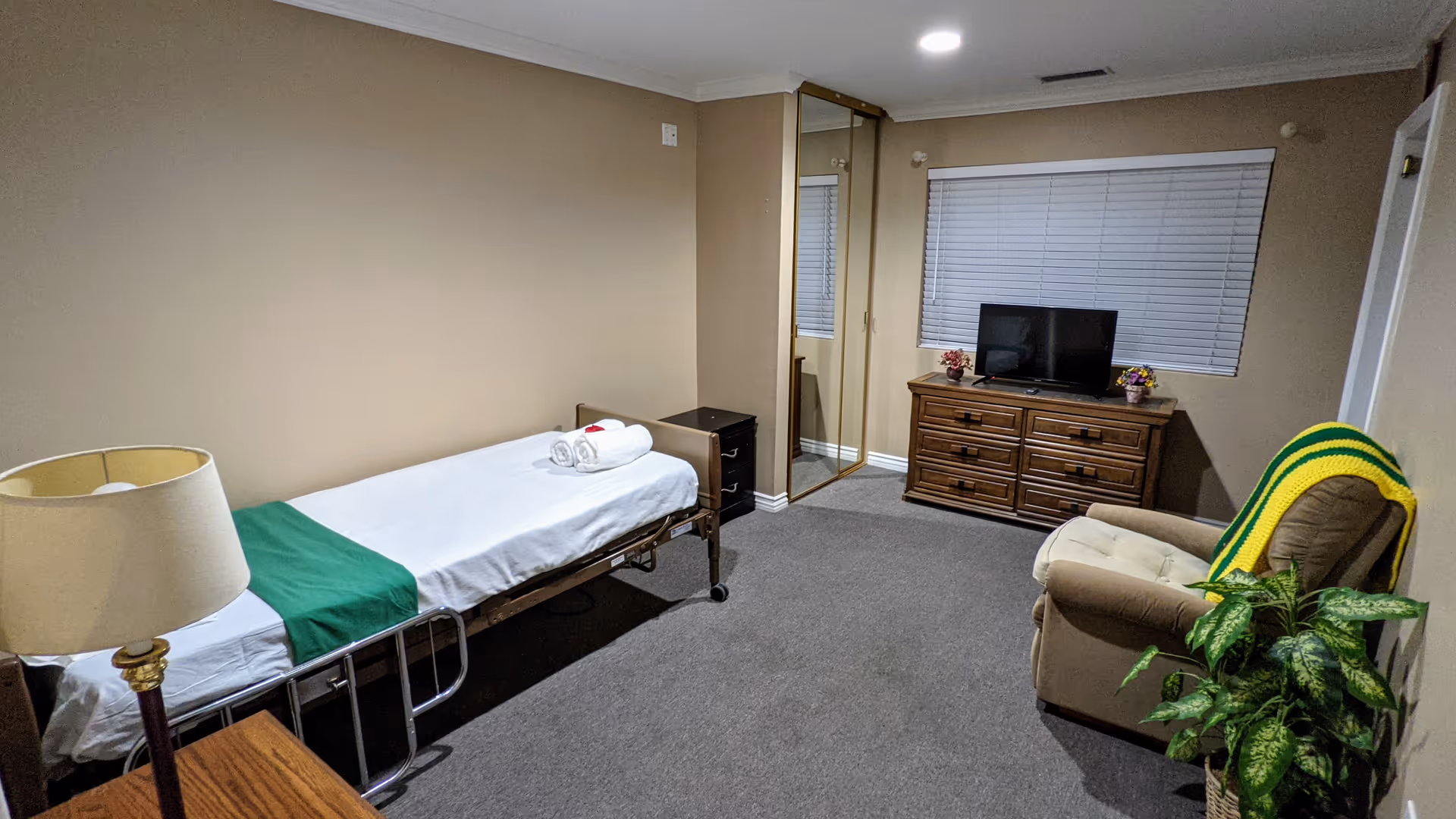 A simple assisted living bedroom with a single bed covered with white sheets and a green blanket at the foot. There is a wooden nightstand with a lamp next to the bed. Across from the bed is a wooden dresser with a flat-screen TV on top, and a window with closed blinds above it. A beige armchair with a green and yellow blanket draped over the back is positioned near a green potted plant. The room has beige walls, carpeted floor, and a mirrored closet door.