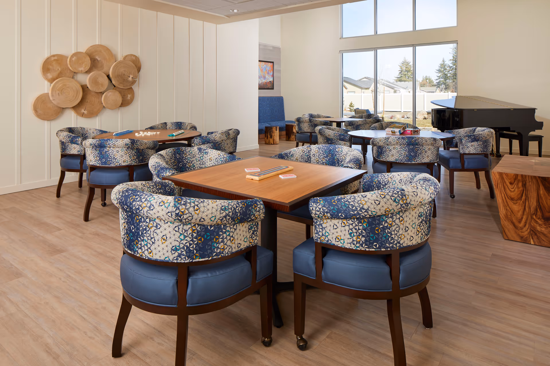Bright communal dining/activity room with patterned upholstered chairs around wooden tables and a grand piano by large windows.