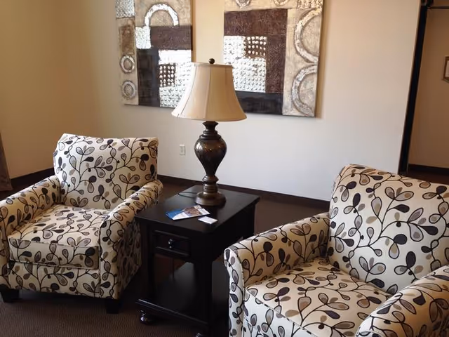 Two patterned armchairs with a floral design are positioned on either side of a dark wooden side table with a decorative lamp on top. Behind the chairs, there is a wall with two abstract art pieces featuring neutral tones.
