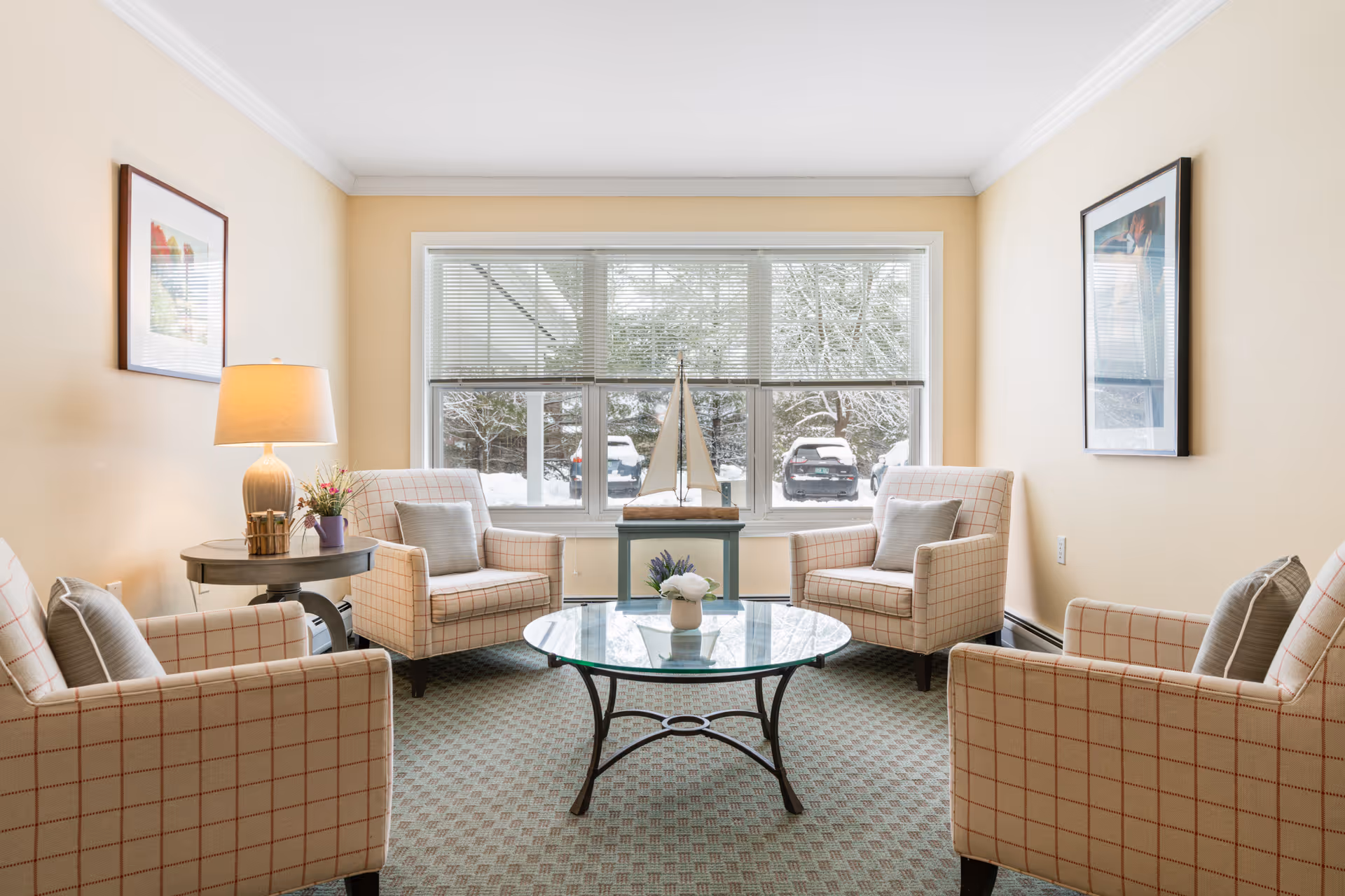Bright common living room with four plaid armchairs arranged around a glass coffee table facing a large window showing snowy outdoors.