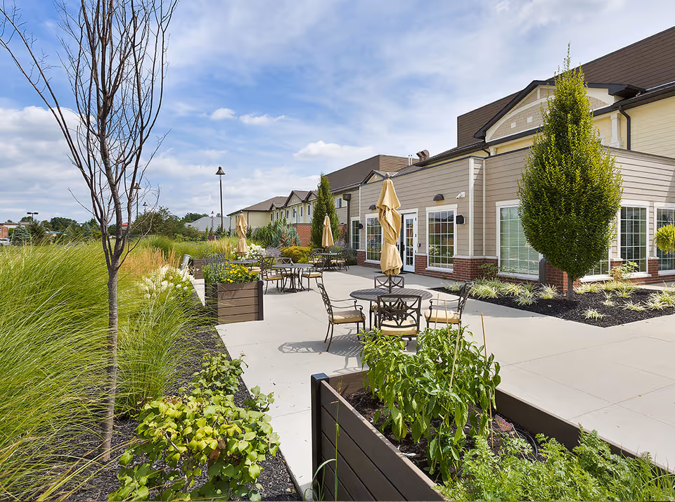 Sunny outdoor patio with round tables and chairs, closed umbrellas, planter boxes, and landscaped greenery beside a two-story building.