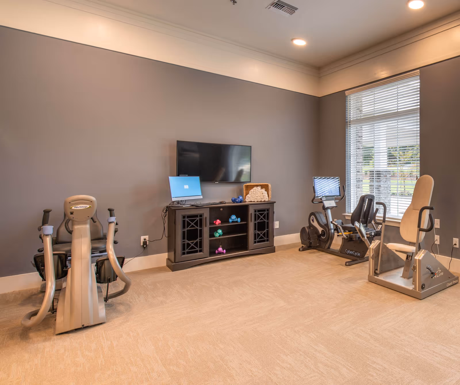 Small fitness room with recumbent exercise bikes, a wall-mounted TV, shelving with hand weights, and a window.