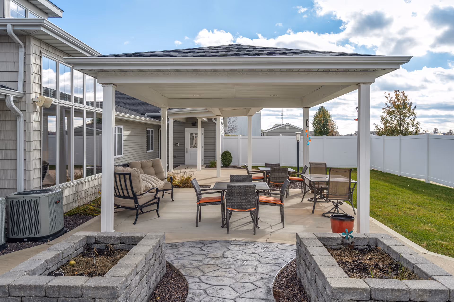 Outdoor covered patio area with several chairs and tables, cushioned seating, and raised garden beds on either side of a stone pathway. The area is adjacent to a building with large windows and a white fence enclosing the grassy yard.