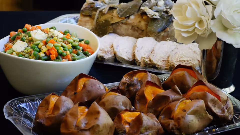 A close-up view of a meal consisting of a bowl of mixed vegetables with butter on top, a platter of sliced roasted meat garnished with herbs, and a tray of baked sweet potatoes. There is also a silver vase with white flowers in the background.