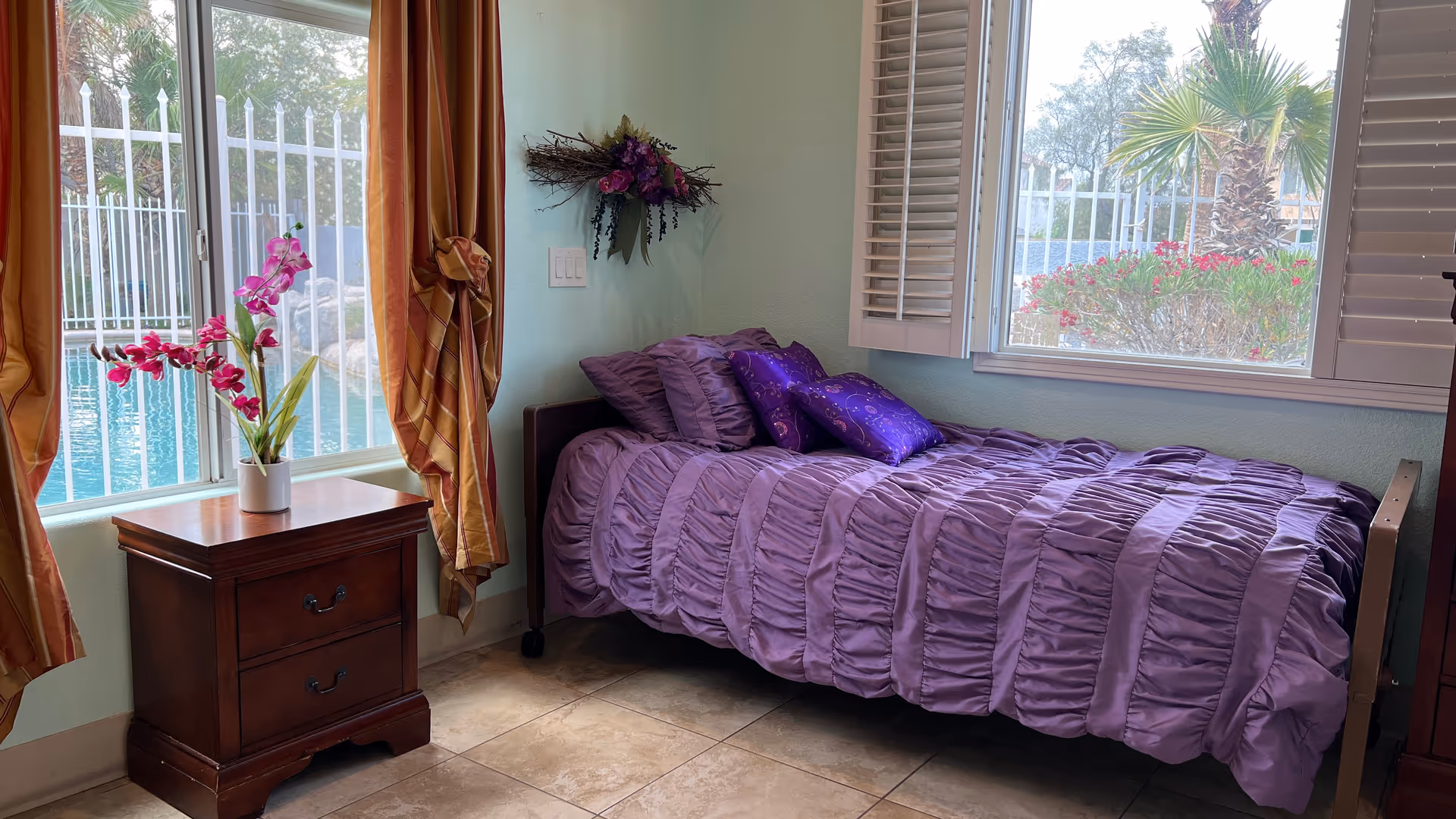 A cozy bedroom with a single bed covered in a purple ruffled comforter and matching pillows. Next to the bed is a wooden nightstand with a vase of pink flowers. The room has two large windows with white shutters and orange curtains, showing a view of a fenced pool and palm trees outside. A decorative floral wreath hangs on the light green wall above the bed.