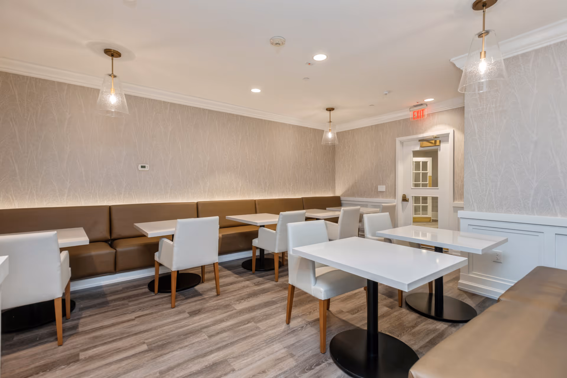 A modern dining room with white tables and white cushioned chairs with wooden legs. There is a long brown cushioned bench along the wall. The room has light-colored wallpaper with a subtle tree branch pattern, wood-style flooring, and three pendant lights hanging from the ceiling. A white door with glass panels and an exit sign above it is visible in the background.