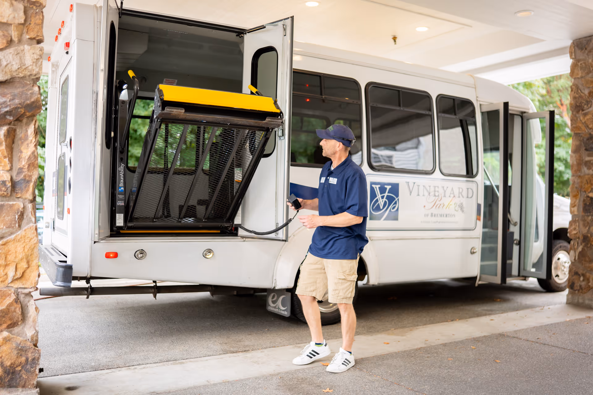 A staff member stands beside a Vineyard Park at Bremerton shuttle bus with its wheelchair lift extended under a covered entrance.