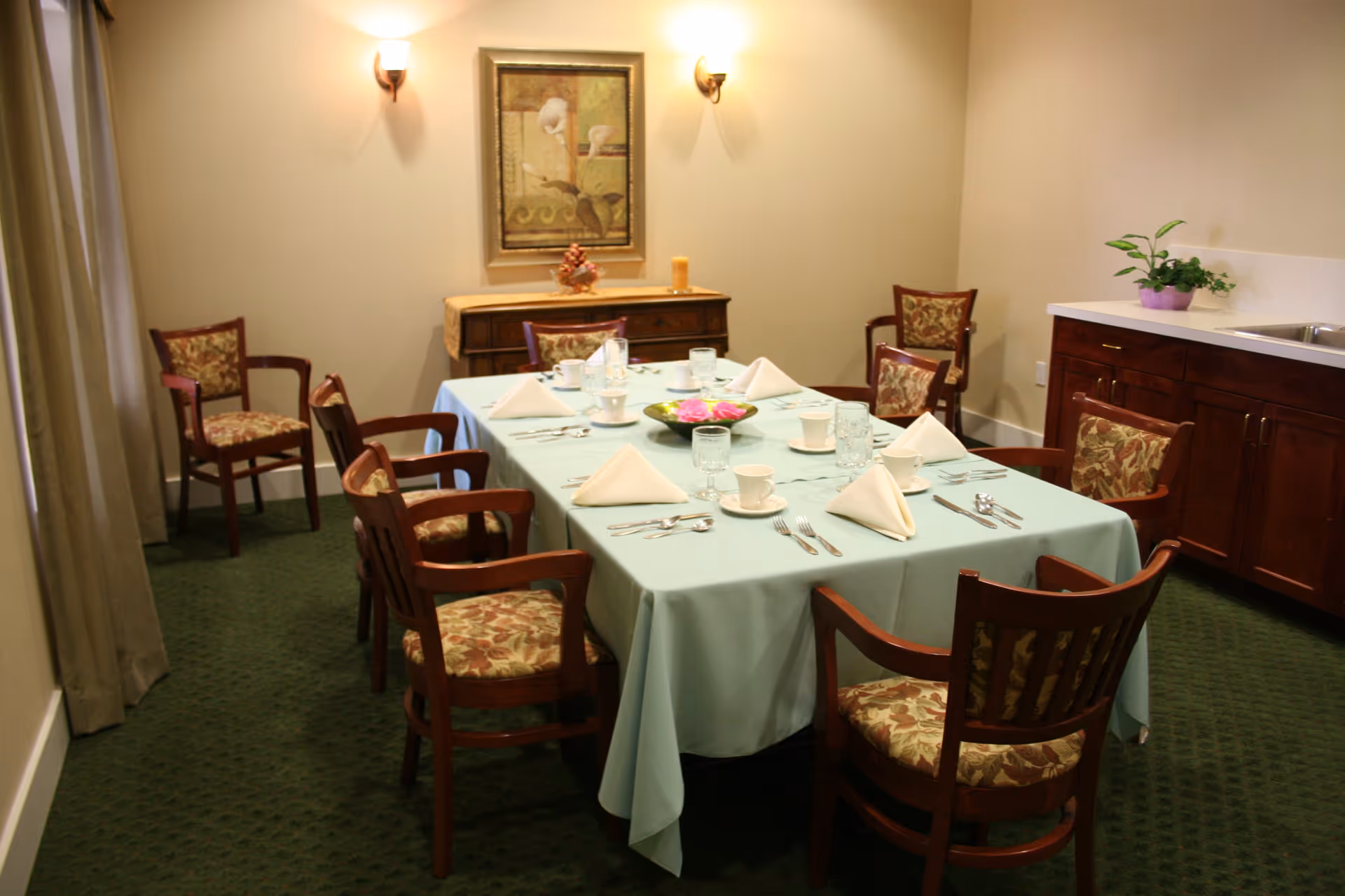 A dining room with a rectangular table covered with a light blue tablecloth. The table is set with white napkins, cups, glasses, and silverware. There are eight wooden chairs with floral-patterned cushions around the table. The room has green carpet, beige walls, a sideboard with a decorative item, a framed painting, two wall sconces, and a countertop with a sink and a small potted plant.