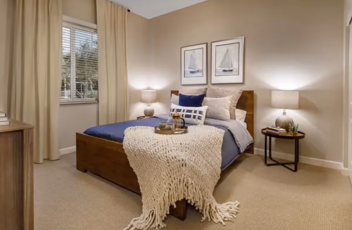 A cozy bedroom with a wooden bed frame, blue bedding, and multiple pillows. A cream-colored knitted throw blanket is draped over the foot of the bed. There are two framed pictures of sailboats on the wall above the bed. Two bedside tables each hold a lamp, with one table also having a small plant and books. A window with beige curtains is on the left side of the room.