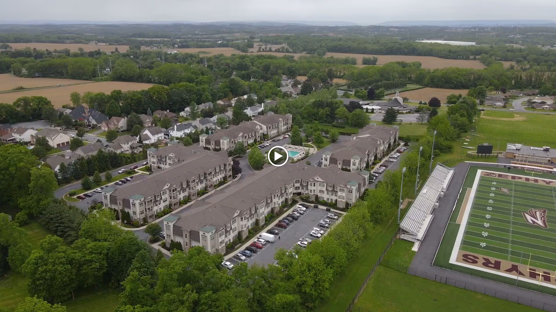 Aerial view of Horizons at the Village at Whitehall, showing multiple residential buildings surrounded by trees and parking lots. Nearby is a sports field with bleachers and a scoreboard. The area is set in a green, suburban landscape with fields and houses in the background.