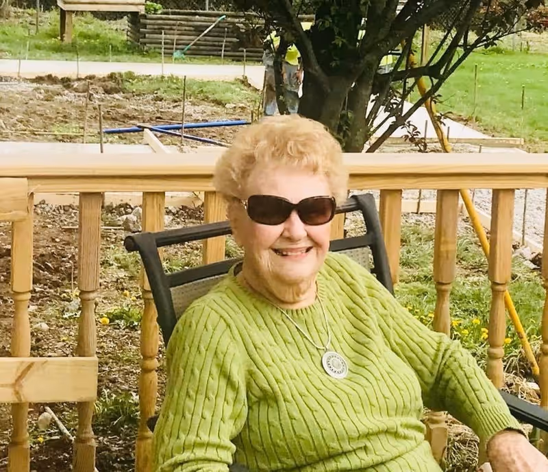 An elderly woman wearing sunglasses and a green sweater is sitting on a chair outdoors on a wooden deck with a garden and tree in the background. She is smiling and appears to be enjoying the sunny day.