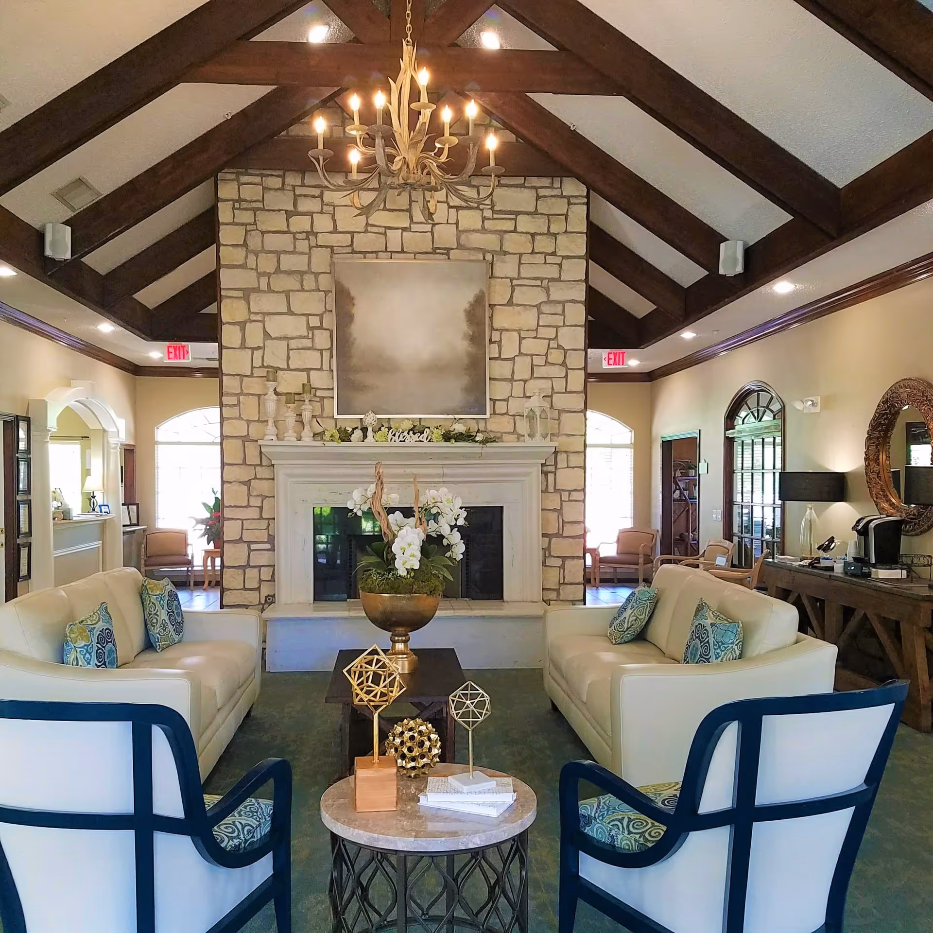 A cozy living room with two white sofas facing each other and two armchairs in the foreground. A stone fireplace with a mantel decorated with candles and a 'blessed' sign is centered on the far wall. Above the fireplace hangs a large, muted landscape painting. A chandelier with multiple lights hangs from the ceiling with exposed wooden beams. There are side tables with decorative objects and a coffee machine on a wooden console table to the right.