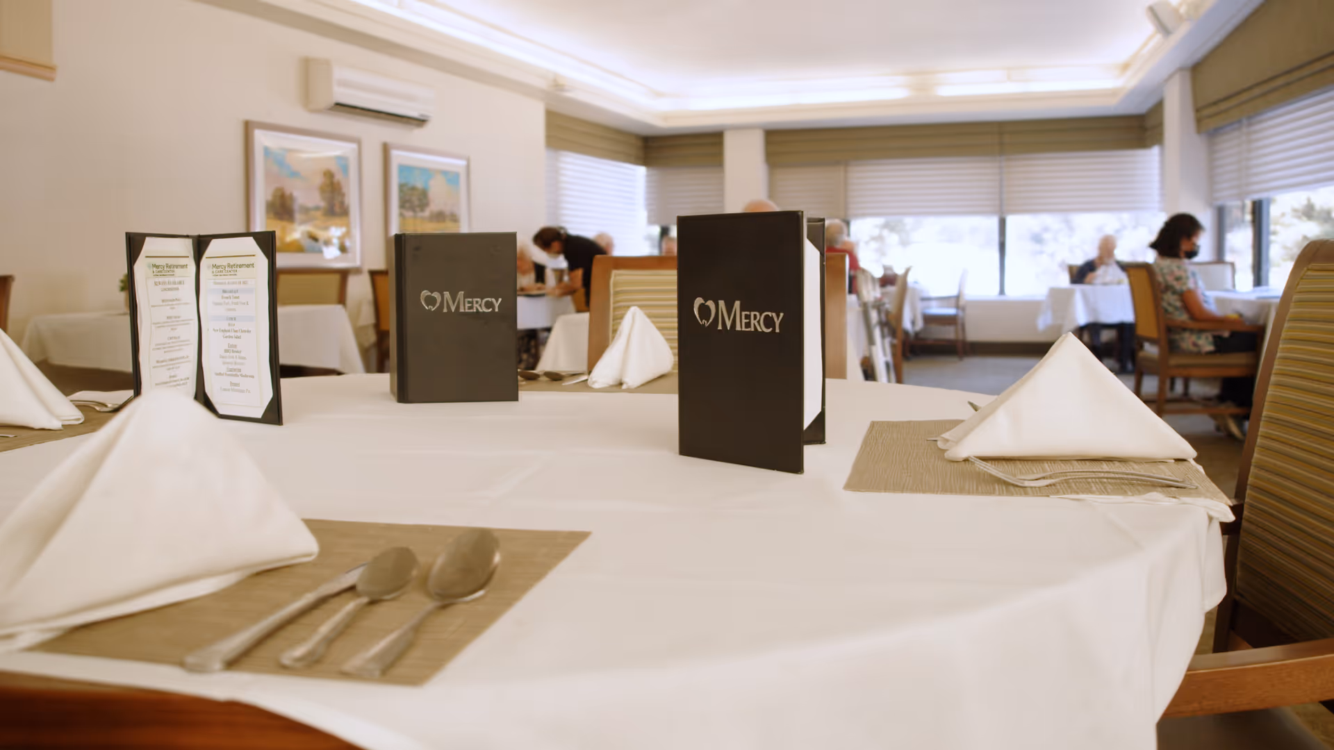 A dining room in Mercy Retirement & Care Center with tables set with white tablecloths, folded white napkins, silverware, and menus. Several elderly people are seated at tables in the background, and a staff member is attending to one of them. The room has large windows with blinds and framed artwork on the walls.