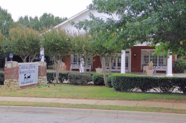 Exterior view of Carriage House Assisted Living of Denton showing a single-story building with a white porch, white columns, and benches. There are trees and bushes in front of the building, along with a sign displaying the facility's name and phone number.