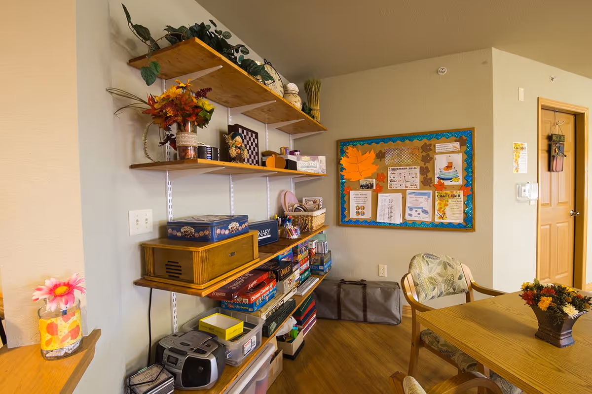 A cozy room with wooden shelves filled with board games, decorative items, and a small stereo. There is a bulletin board on the wall with various notices and a large orange leaf decoration. A wooden table with a floral centerpiece and cushioned chairs is partially visible. The room has light-colored walls and a wooden door in the background.