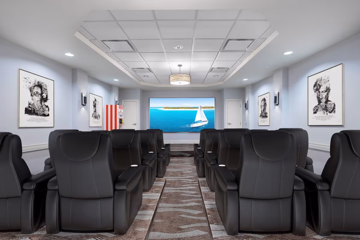 A modern theater room with two rows of black leather recliner chairs facing a large screen displaying a sailboat on blue water. The room has light gray walls adorned with framed black and white artistic portraits, recessed ceiling lights, and a patterned carpet. A popcorn machine is visible on the left side near the back.