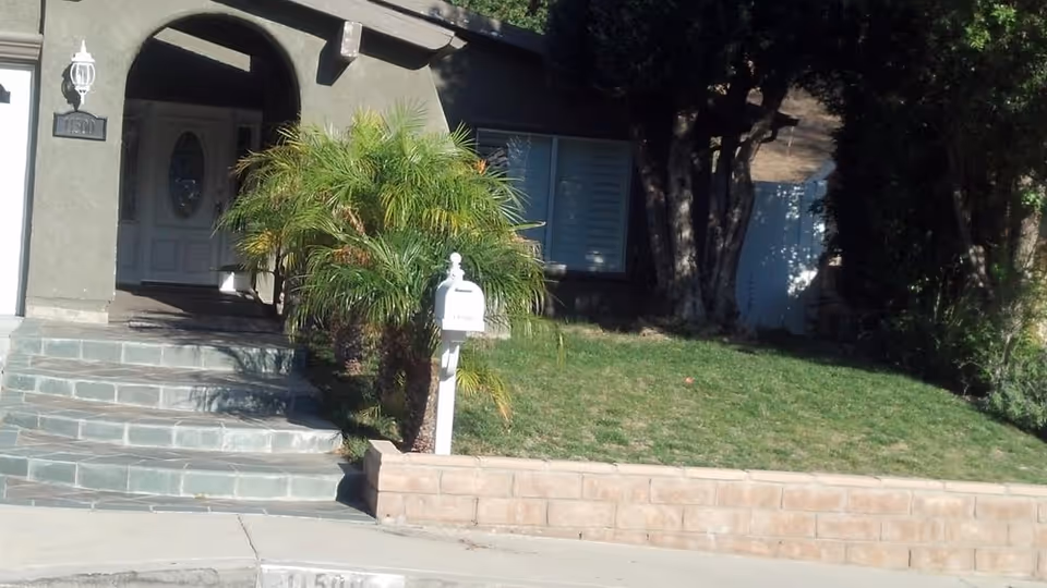 Front exterior view of a residential building with a stone staircase leading to a covered entrance. There is a white mailbox on a post near a small palm tree and a grassy lawn bordered by a low brick retaining wall.