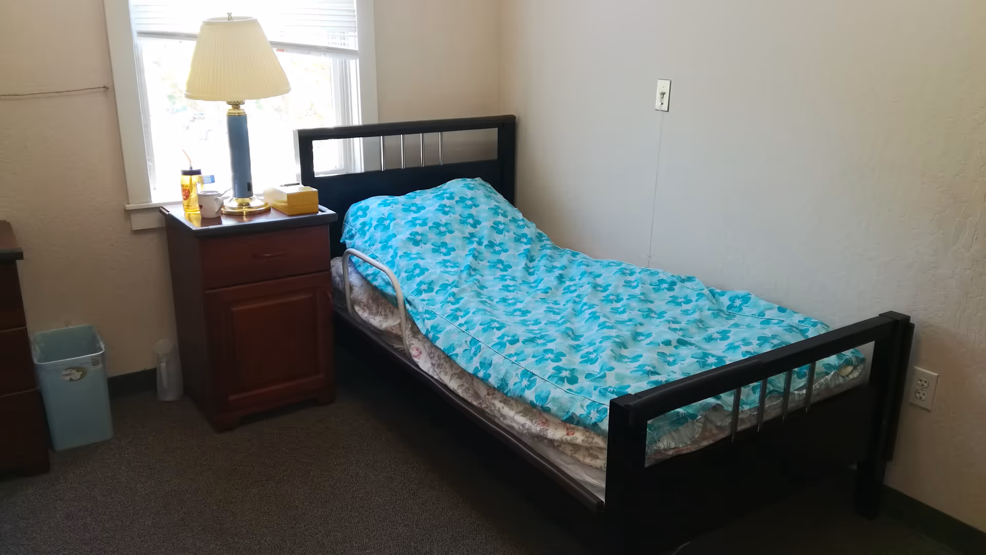 A small bedroom with a single bed covered with a blue floral blanket. Next to the bed is a wooden nightstand with a lamp, a yellow tissue box, a cup, and a yellow water bottle. There is a window behind the nightstand letting in natural light. A small trash bin is on the floor near the nightstand.