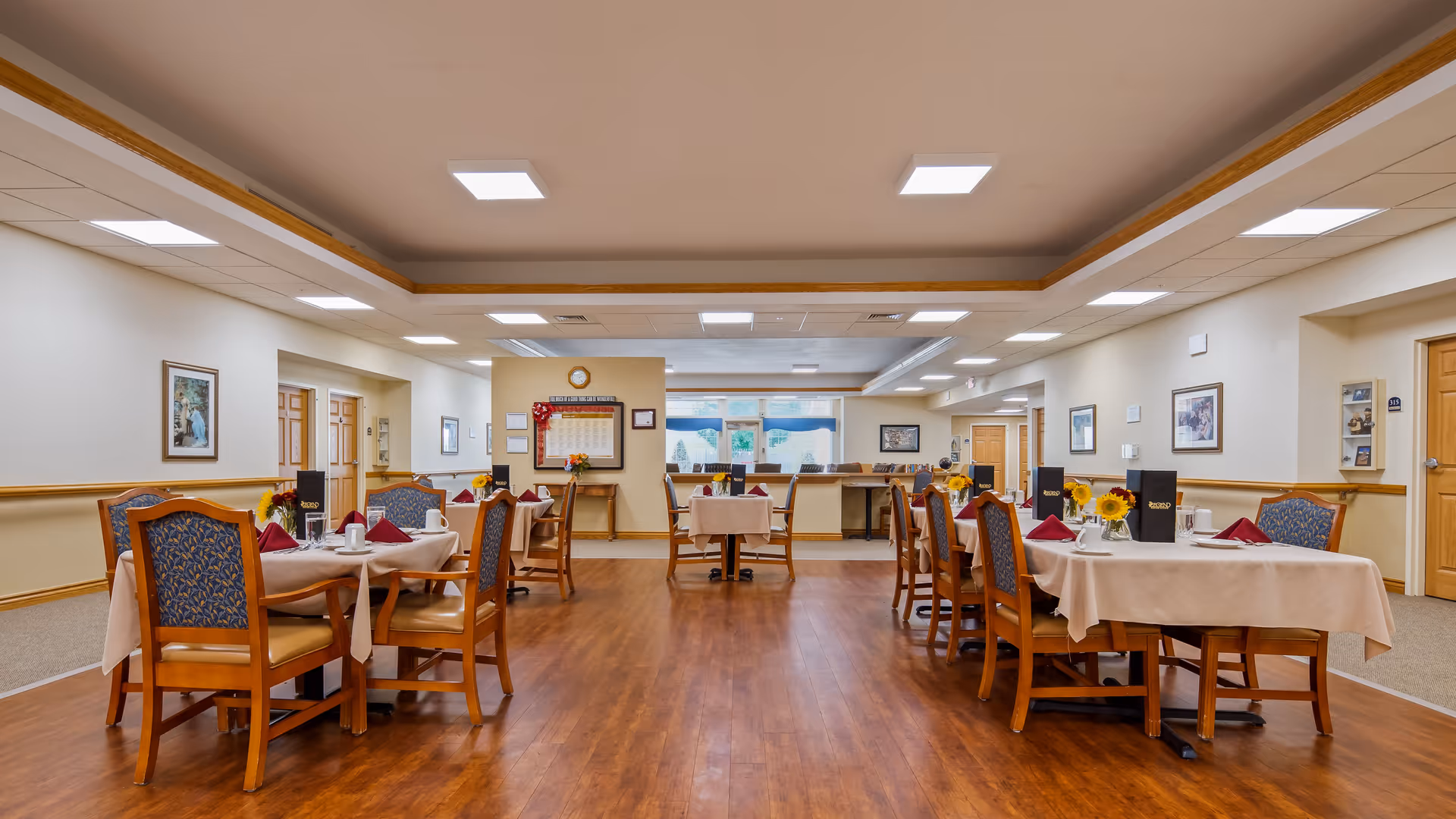 A spacious dining room in a senior living facility with several tables covered in beige tablecloths, each set with plates, cups, napkins, and menus. The room has wooden flooring, cushioned chairs with patterned backs, framed artwork on the walls, and bright ceiling lights.