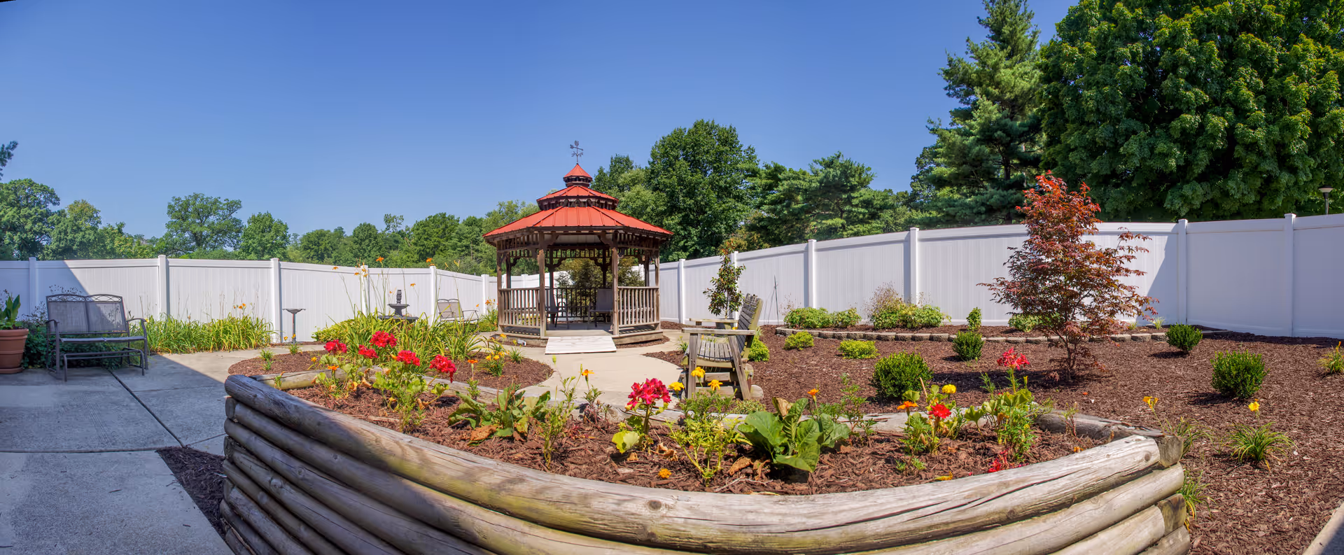 Outdoor garden area with a wooden gazebo in the center, surrounded by flower beds with red and yellow flowers, small shrubs, and a white privacy fence. There are benches and chairs placed around the garden, and tall green trees in the background under a clear blue sky.