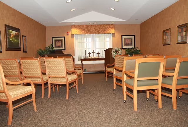 A small chapel or meditation room with rows of wooden chairs facing a table with three crosses and a lectern. The room has beige walls with framed artwork, a window with a valance, and carpeted floor.