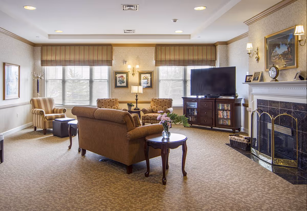 A cozy living room with beige carpet and neutral-colored walls featuring a fireplace with a decorative screen and mantel adorned with a clock and framed pictures. The room has a large flat-screen TV on a wooden cabinet, two patterned armchairs, a striped armchair, a brown sofa, and a round wooden side table with a vase of flowers. Large windows with striped valances allow natural light to fill the space.