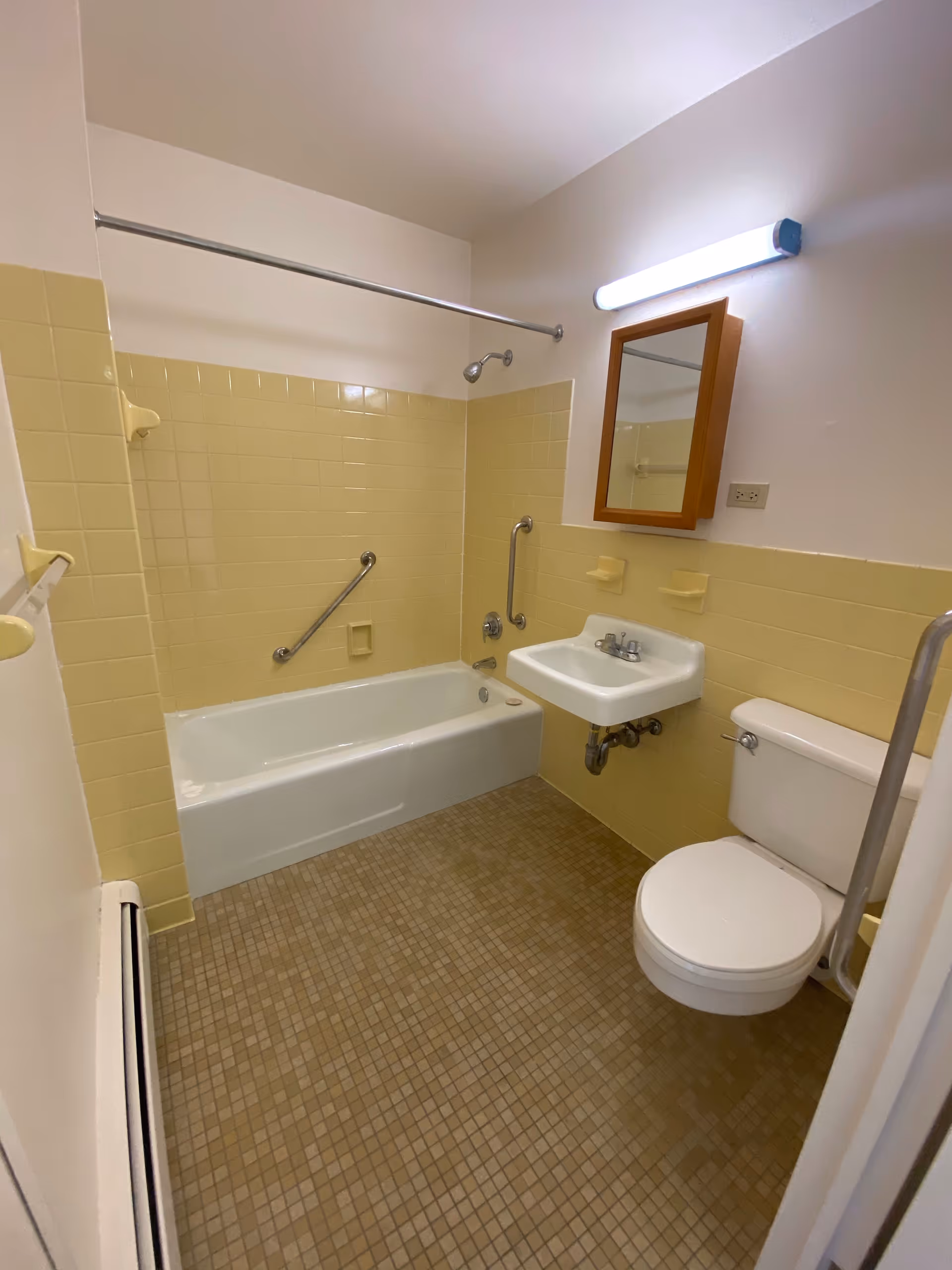 Small accessible bathroom with a bathtub, wall-mounted sink, toilet, grab bars, and yellow tile walls.
