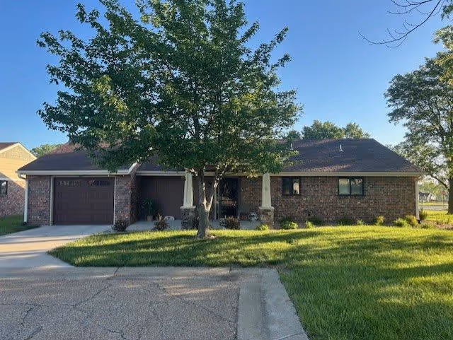 Single-story brick house with a dark roof, a garage on the left, a tree in the front yard, and a well-maintained lawn under a clear blue sky.