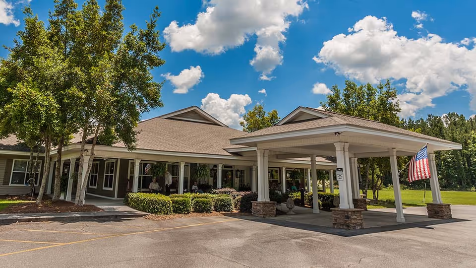 Exterior view of Fellowship Nashville Assisted Living Community building with a covered entrance supported by white columns, an American flag hanging from the right side, surrounded by trees and bushes under a partly cloudy blue sky.