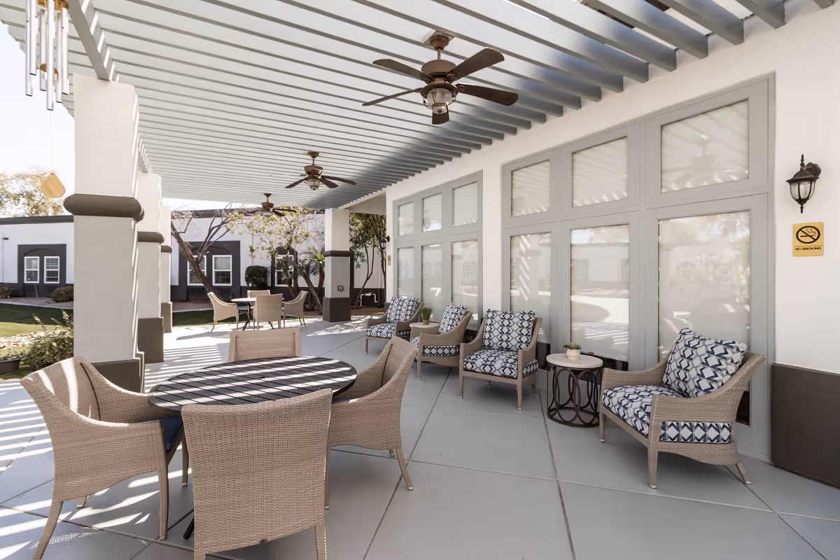 Outdoor covered patio area with wicker chairs and tables, ceiling fans, and large windows. The space is furnished with cushioned armchairs and round tables, surrounded by a garden and other buildings in the background.