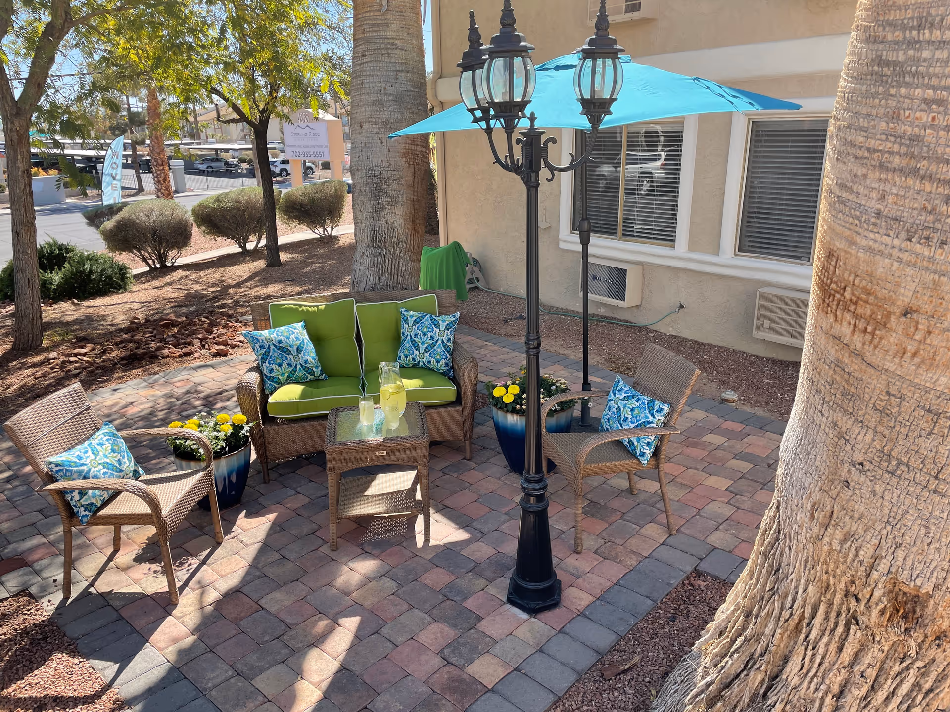 Outdoor patio area with wicker furniture including a loveseat and two chairs with green and blue patterned cushions. A small glass-top table holds a pitcher of lemonade and two glasses. A black vintage-style street lamp with a blue umbrella attached provides shade. The patio is surrounded by trees, bushes, and a building with windows in the background.