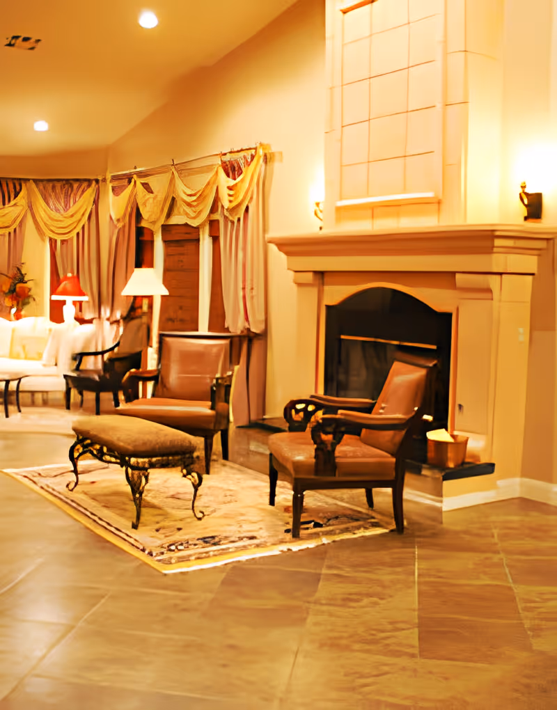 A cozy living room area with two brown armchairs and a matching ottoman placed on a decorative rug in front of a large beige fireplace. The room features warm lighting, elegant curtains with valances, and additional seating including a white sofa and black chairs in the background.