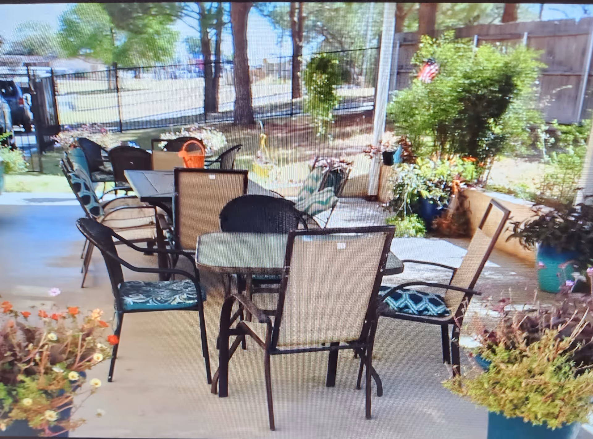 Outdoor patio area with multiple tables and chairs arranged for seating, surrounded by potted plants and greenery. The area is shaded and enclosed by a fence with trees and a lawn visible in the background.