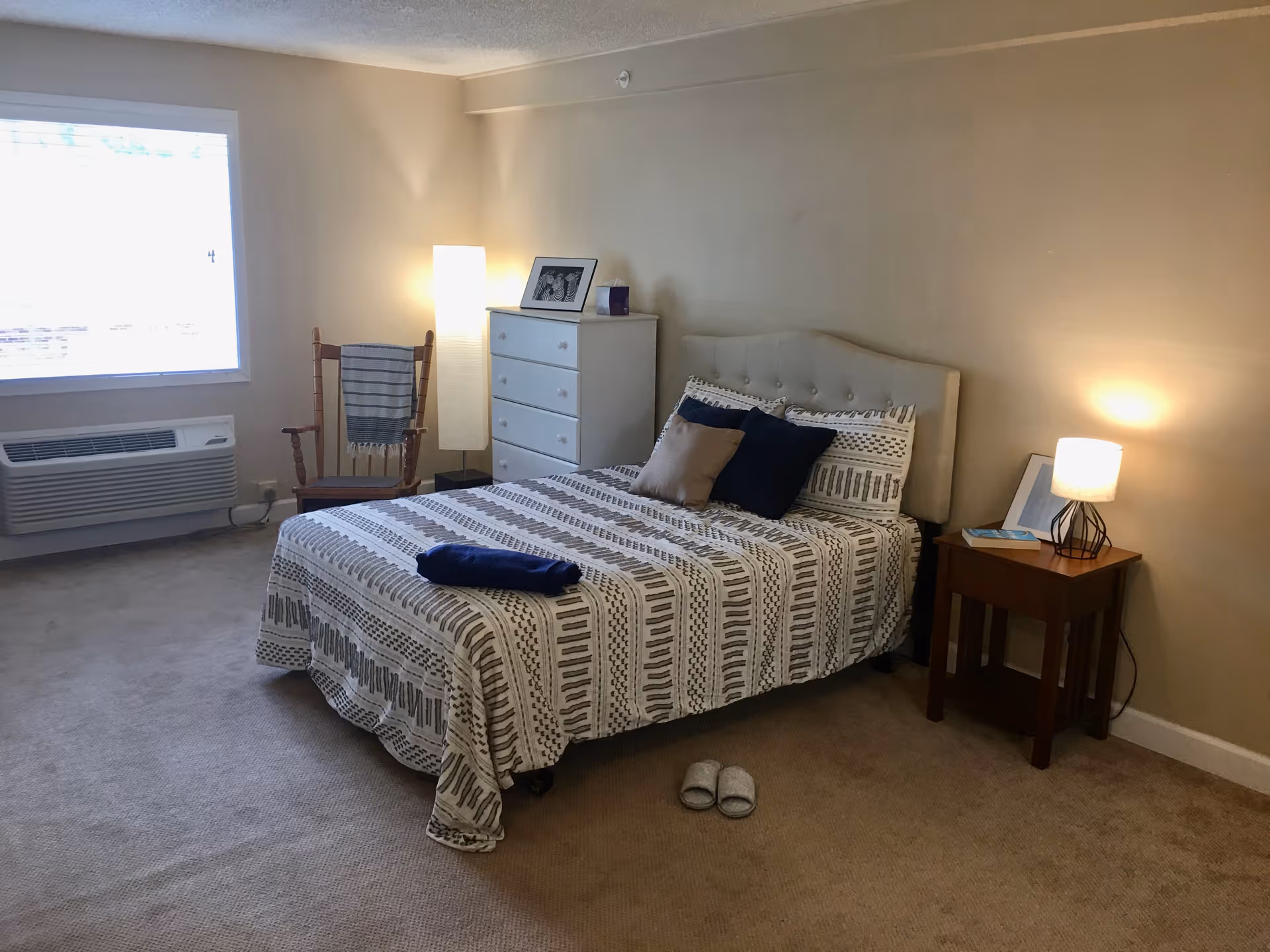 A cozy bedroom with a bed covered in a patterned bedspread and several pillows. There is a wooden nightstand with a lamp and a book, a white chest of drawers with a framed photo and a candle on top, a wooden chair with a striped blanket draped over it, and a tall floor lamp. A window with blinds lets in natural light, and a pair of slippers is placed on the carpeted floor near the bed.