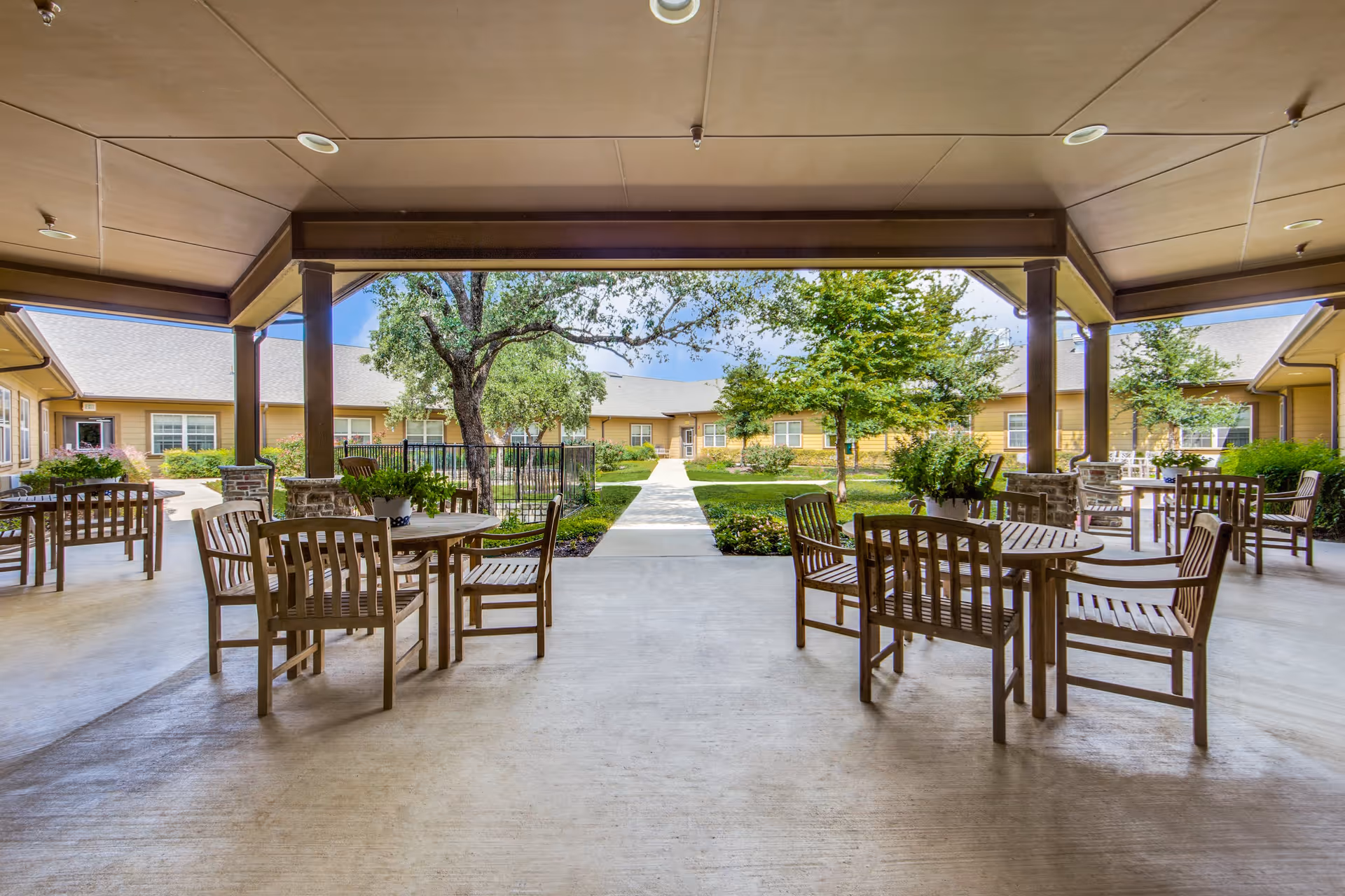 Covered outdoor patio area with several wooden tables and chairs arranged for seating, overlooking a landscaped courtyard with trees, shrubs, and a paved walkway leading to a building with multiple windows under a clear blue sky.