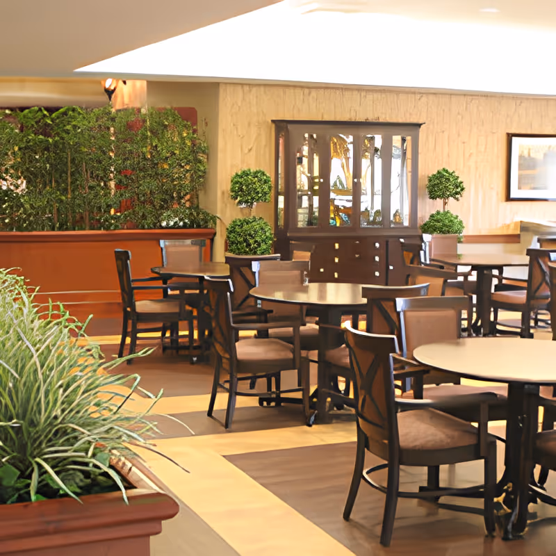 A well-lit dining area with round tables and wooden chairs arranged neatly. The room features decorative plants in planters and topiary, a wooden cabinet with glass doors, and framed artwork on the wall. The flooring has a combination of wood and tile patterns.