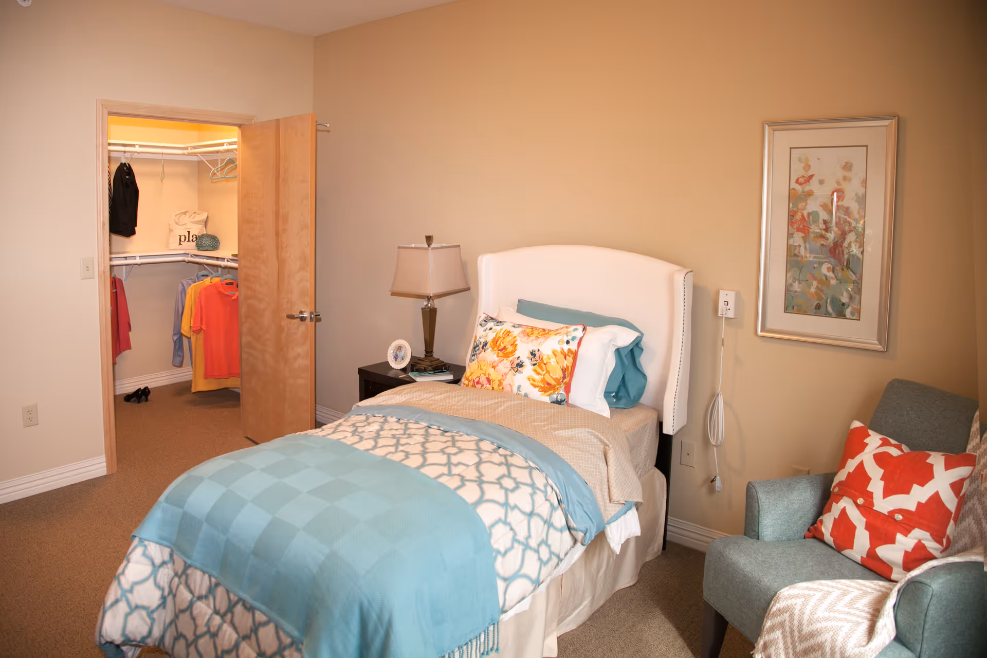 A cozy bedroom with a single bed dressed in patterned and solid blue bedding, a floral pillow, and a beige blanket. Next to the bed is a dark wooden nightstand with a lamp and a small clock. An armchair with a red and white patterned pillow and a beige throw blanket is positioned to the right. A framed abstract floral painting hangs on the wall above the chair. The door to a walk-in closet is open, revealing hanging clothes and shelves inside.