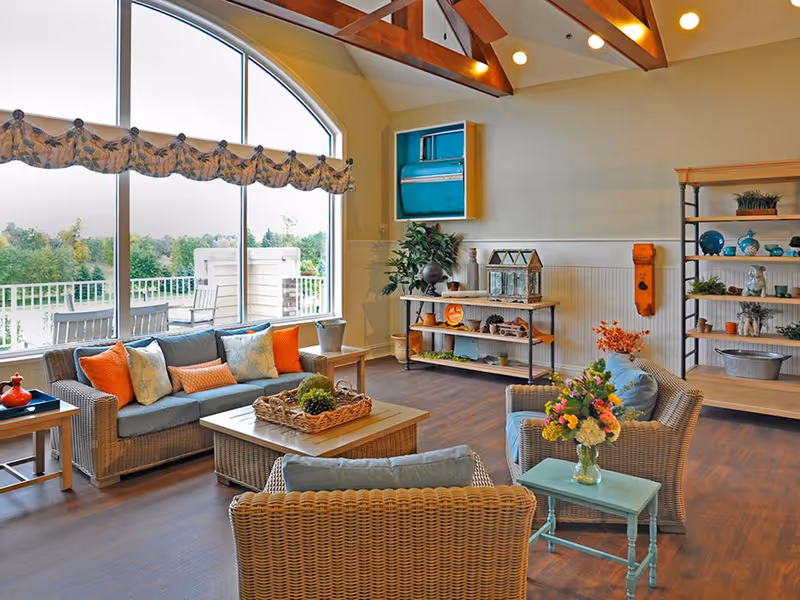 A bright and cozy living room in a senior living facility with large arched windows letting in natural light. The room features wicker furniture including a sofa and chairs with blue and orange cushions, a coffee table with a decorative basket, and side tables. There are shelves with decorative items and plants, wooden beams on the ceiling, and a ceiling fan. Outside the window, greenery and outdoor seating are visible.