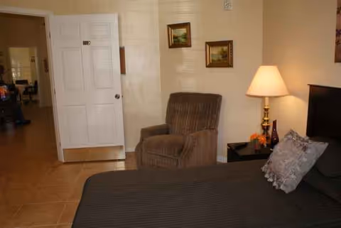 A furnished bedroom in a senior living facility with bed, armchair, lamp, and side table.