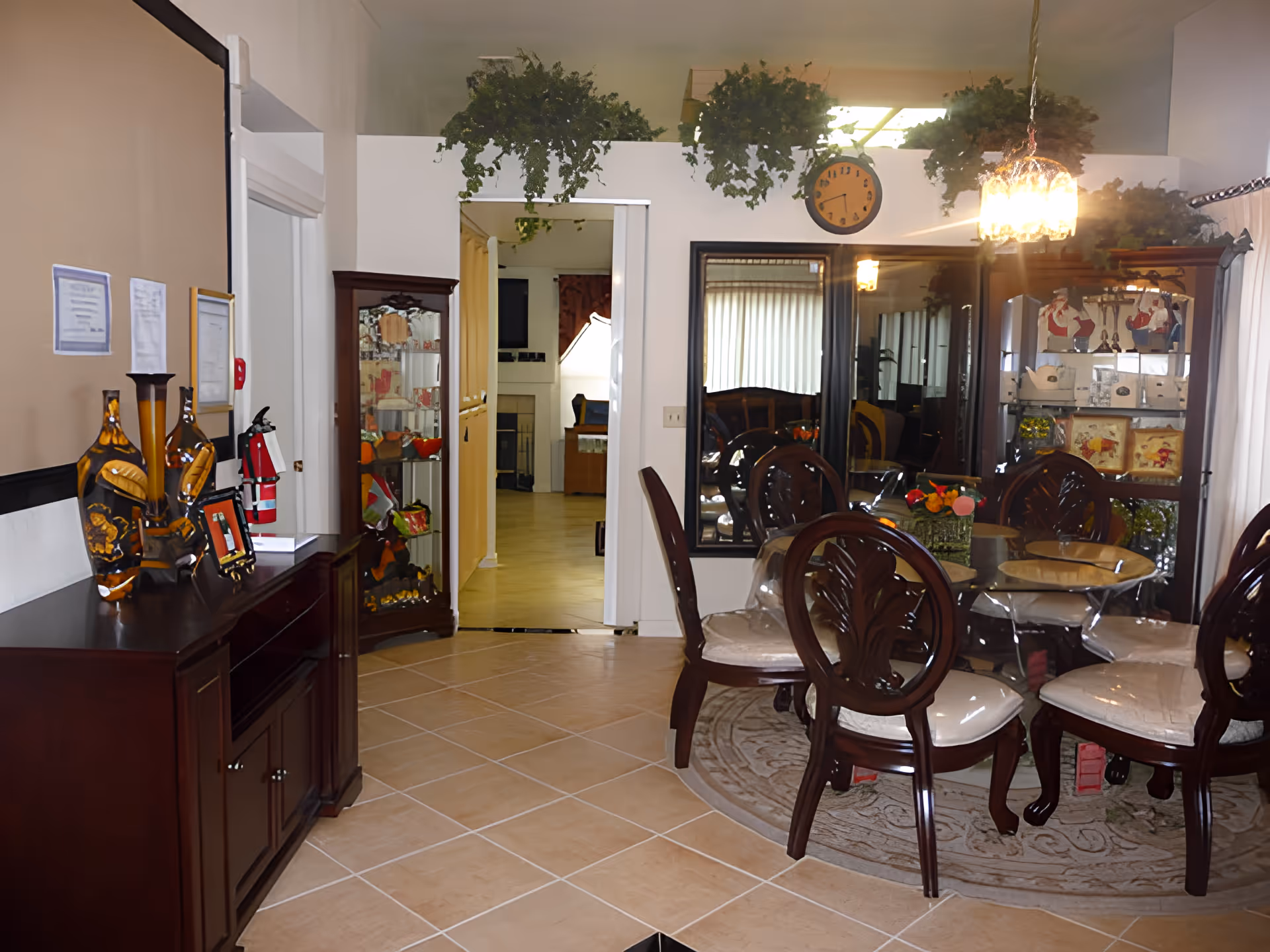 Interior view of a dining area with a round glass table surrounded by six wooden chairs with cushioned seats. The room features a decorative rug under the table, a large mirror on the wall, a hanging light fixture above the table, and a wooden cabinet with decorative items and plants on top. There is an open doorway leading to another room with a fireplace and additional seating.