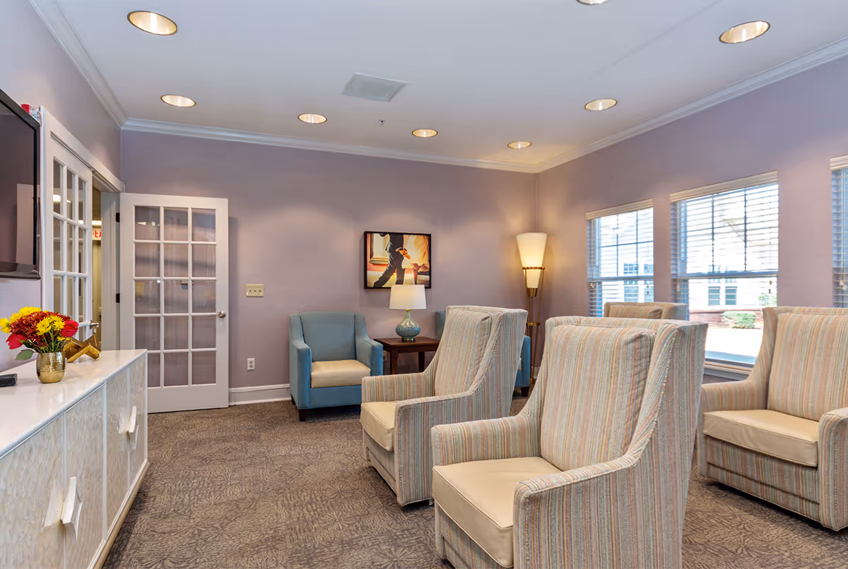 A cozy living room area with several striped armchairs and a blue armchair arranged around a small wooden side table with a lamp. The walls are painted light purple, and there is a framed picture hanging on the wall. Large windows with blinds allow natural light to enter the room. A white cabinet with a vase of flowers and a TV mounted above it is visible on the left side. The room has recessed ceiling lights and a floor lamp in the corner.