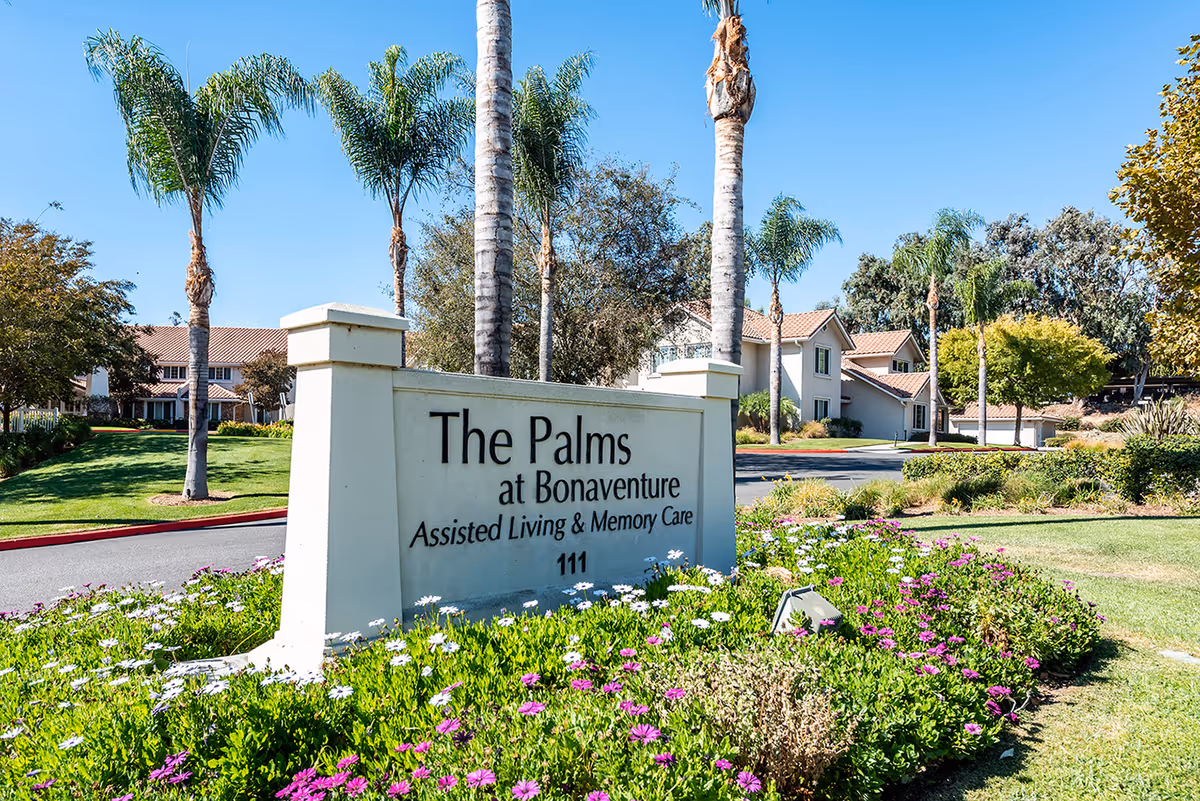Entrance sign for The Palms at Bonaventure Assisted Living & Memory Care surrounded by green grass, colorful flowers, palm trees, and residential buildings in the background under a clear blue sky.