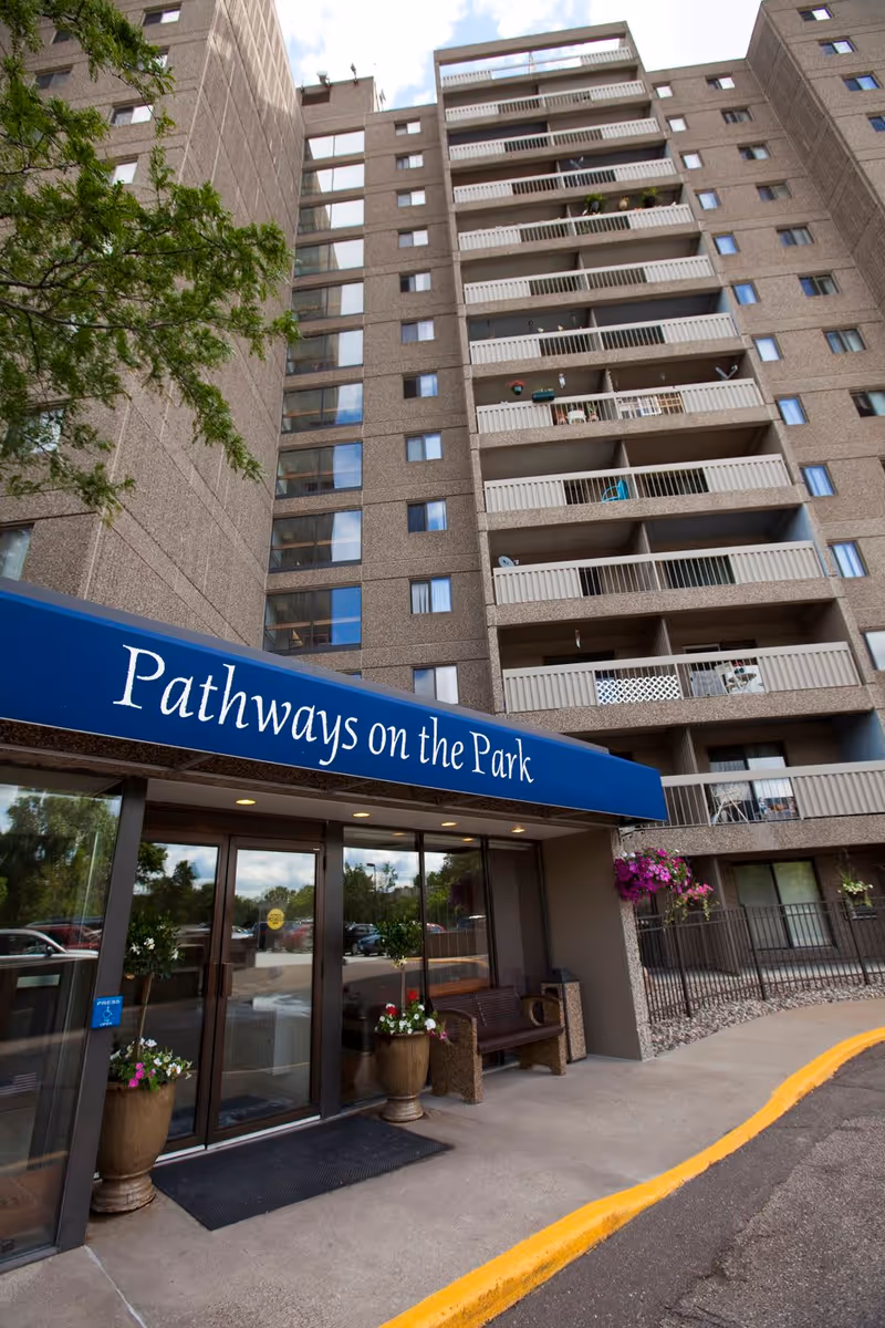 Exterior view of a multi-story residential building with balconies. The entrance has a blue awning with the text 'Pathways on the Park'. There are potted plants and a bench near the entrance, and a tree is partially visible on the left side.