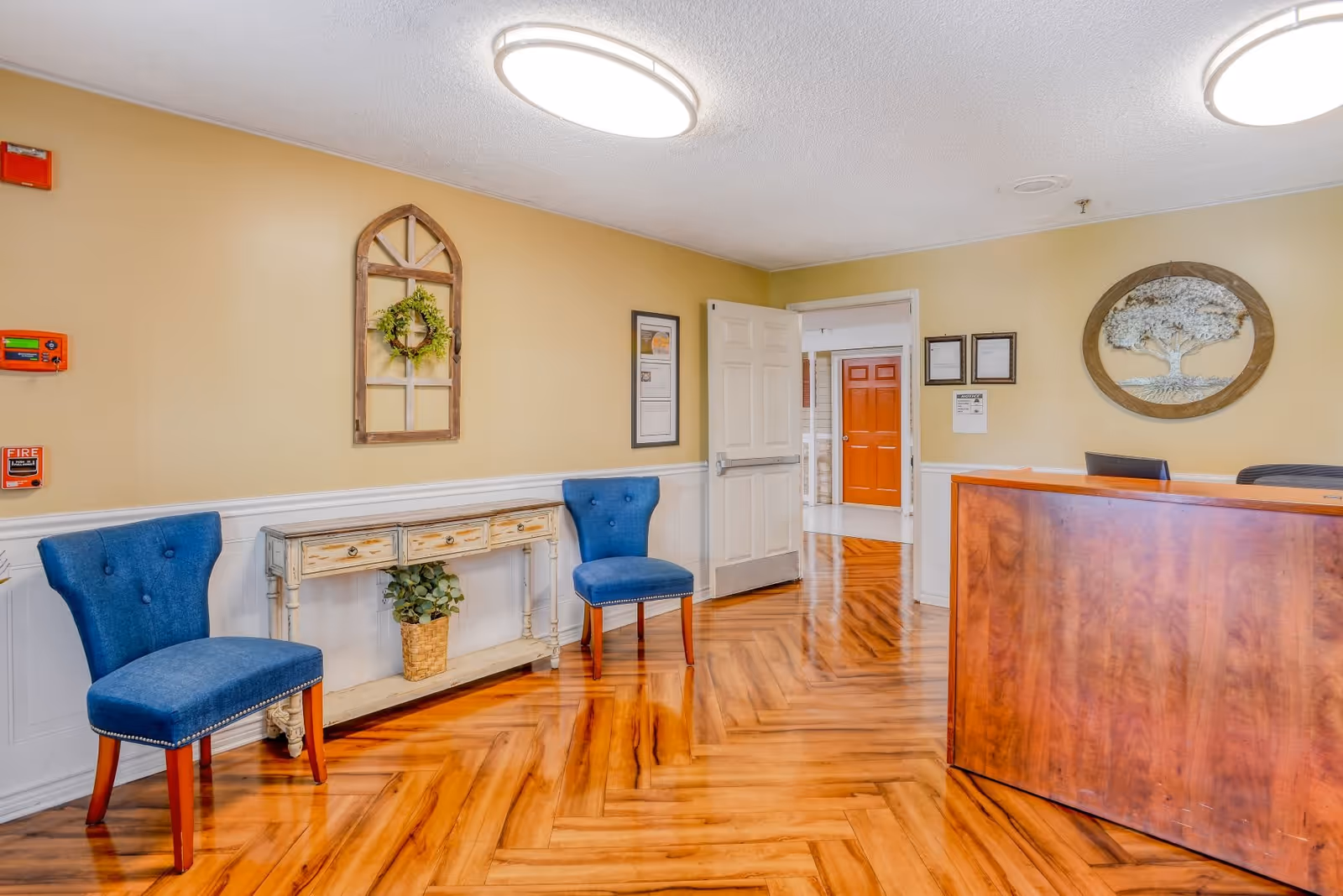 Reception area of Great American Assisted Living at Tampa featuring a wooden reception desk, two blue upholstered chairs, a decorative console table with a plant underneath, wall decorations including a wreath and framed certificates, and a polished wooden floor.