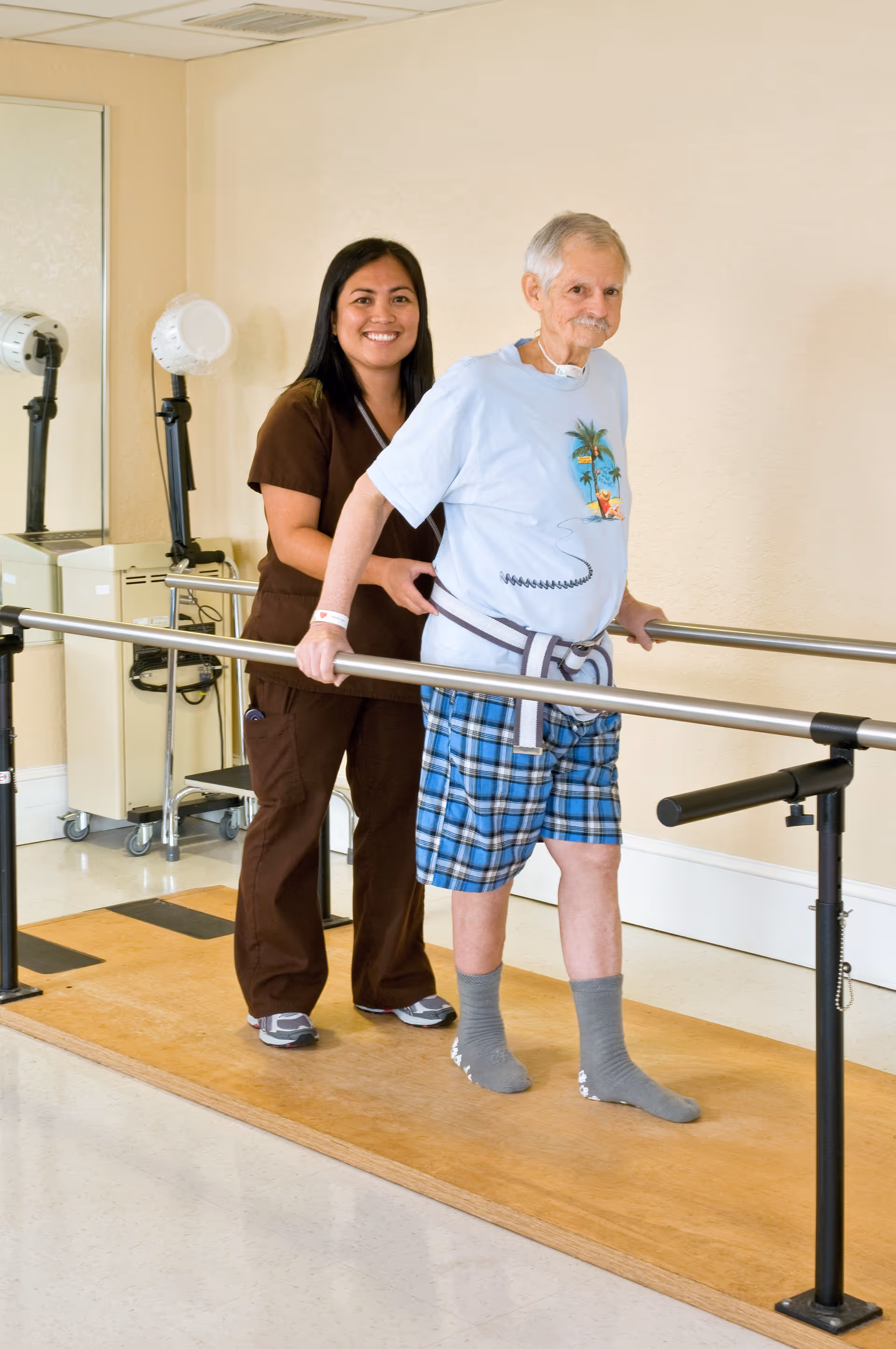 A healthcare worker assists an elderly man with physical therapy as he walks between parallel bars in a rehabilitation room. The man is wearing a light blue t-shirt with a tropical print, plaid shorts, and gray socks, while the healthcare worker is dressed in brown scrubs and smiling.