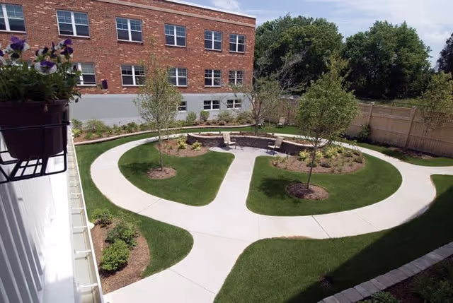 A landscaped outdoor courtyard with a circular concrete walkway surrounding small trees and garden beds. There are a few chairs placed around the inner circle. A brick building with multiple windows is visible in the background, along with a wooden fence and some large trees.