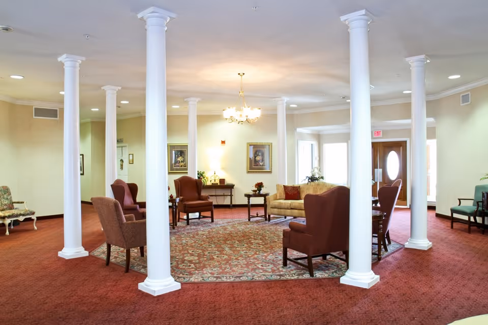 Lobby seating area with white columns, red carpet, patterned rug and arranged armchairs and a sofa under a central chandelier.