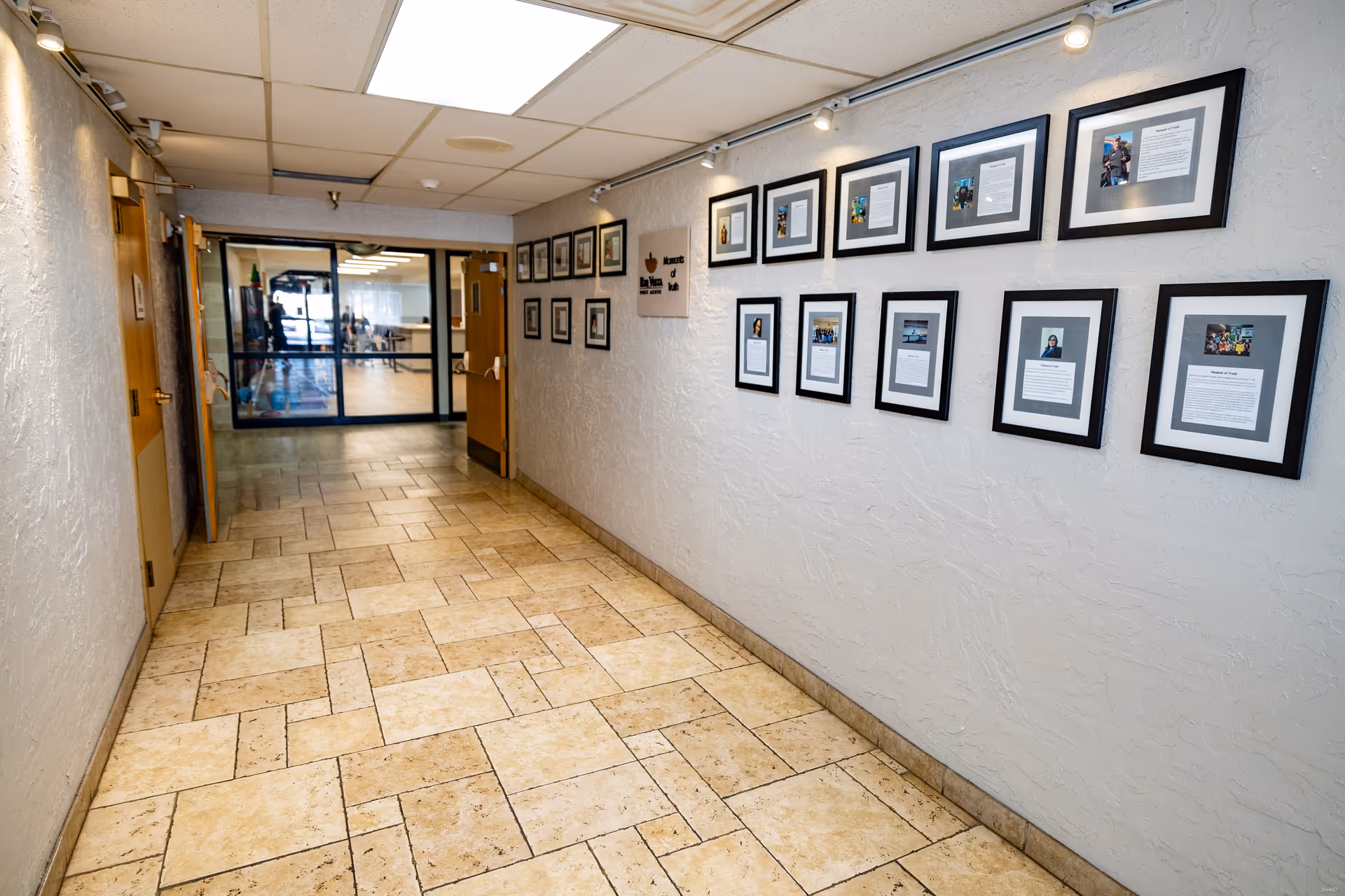 A hallway with beige tiled floor and textured white walls. The right wall is decorated with framed photos and documents arranged in two rows. The hallway leads to a set of double glass doors at the end, with wooden doors on the left side. Ceiling lights illuminate the corridor.