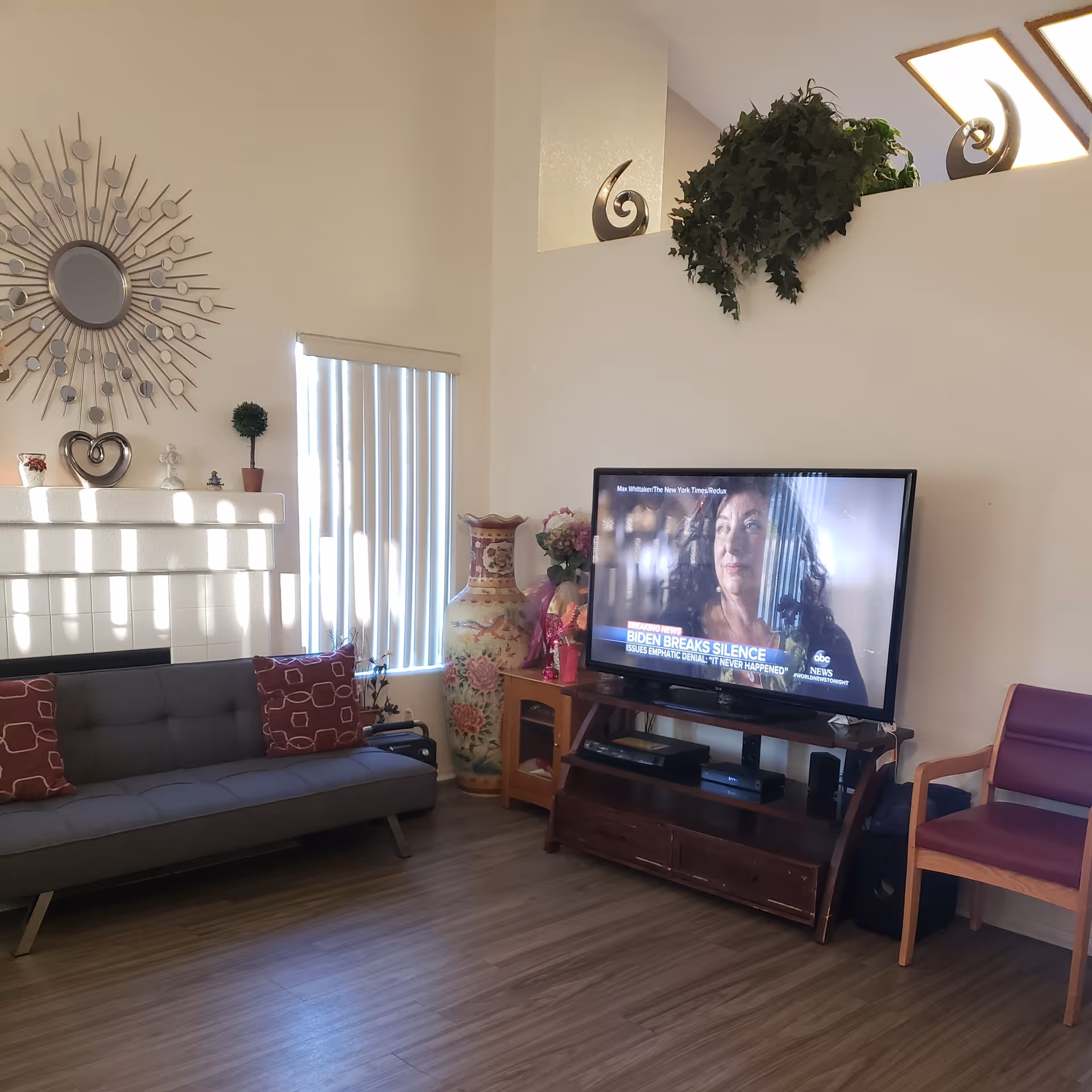 A cozy living room with a gray sofa adorned with red patterned cushions, a large decorative vase with floral designs, a flat-screen TV on a wooden stand showing a news segment, a purple chair with wooden arms, and decorative items on a white mantel and shelf above the TV. The room has wooden flooring and vertical blinds on the window.