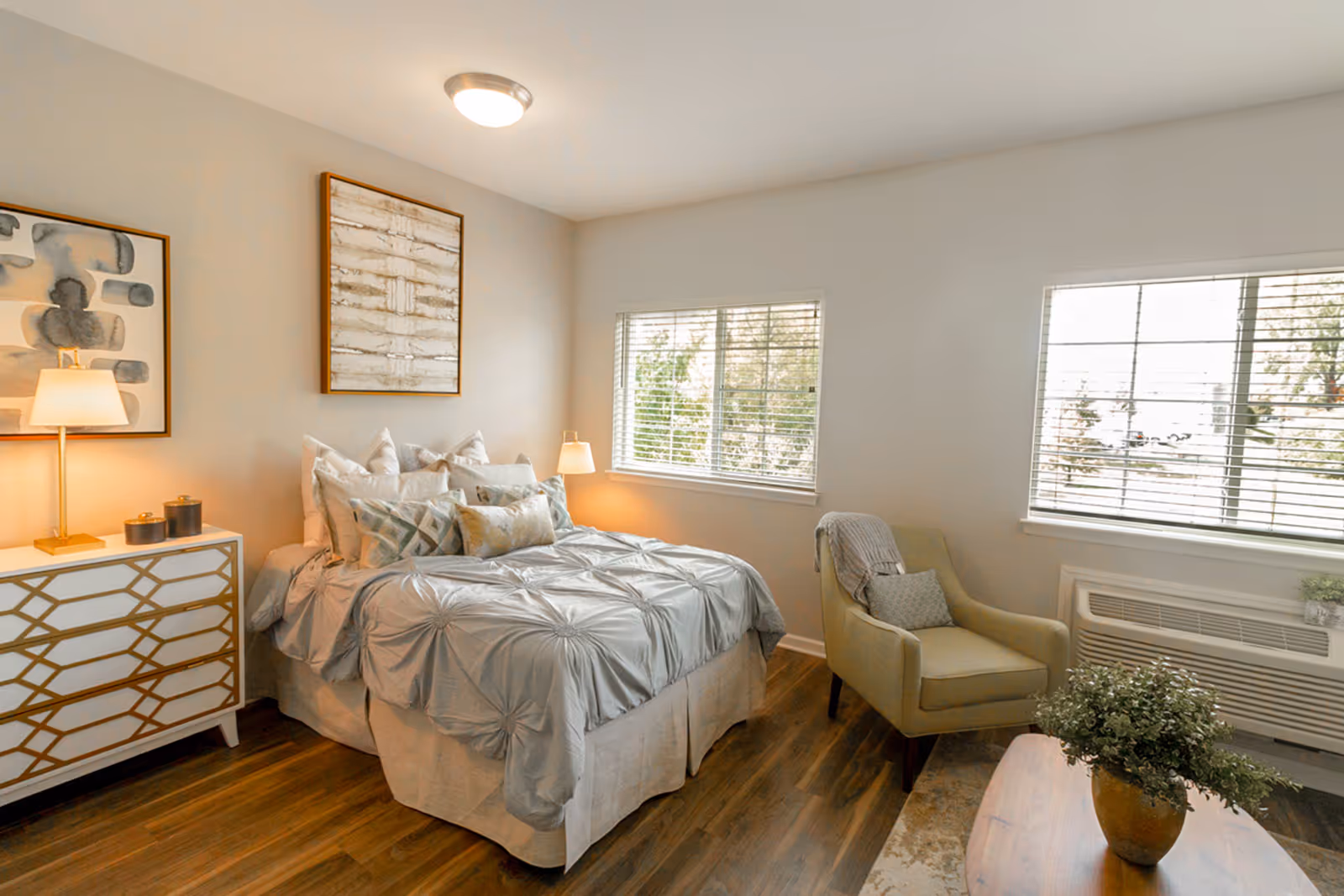 A cozy bedroom with a neatly made bed featuring multiple pillows and a light gray comforter. There is a white dresser with gold geometric patterns on the left side, topped with a lamp and decorative items. Two framed abstract artworks hang on the wall above the bed. To the right, a green upholstered armchair with a throw blanket and pillow sits near two windows with blinds. A wooden coffee table with a potted plant is in the foreground, and the room has wood flooring and neutral-colored walls.